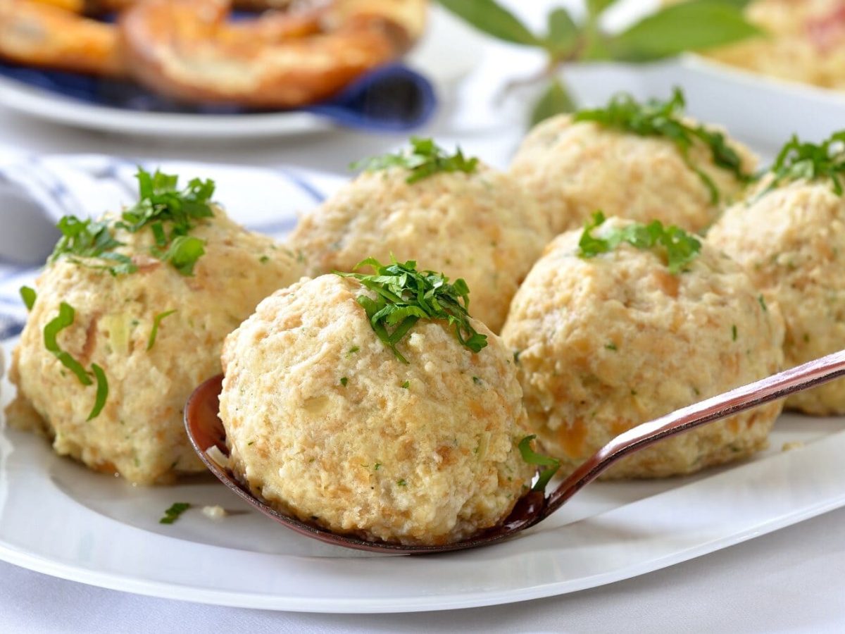 Bayerische Semmelknödel mit Petersilie liegen auf einer weißen Servierplatte, dahinter liegen Brezeln auf einem Teller