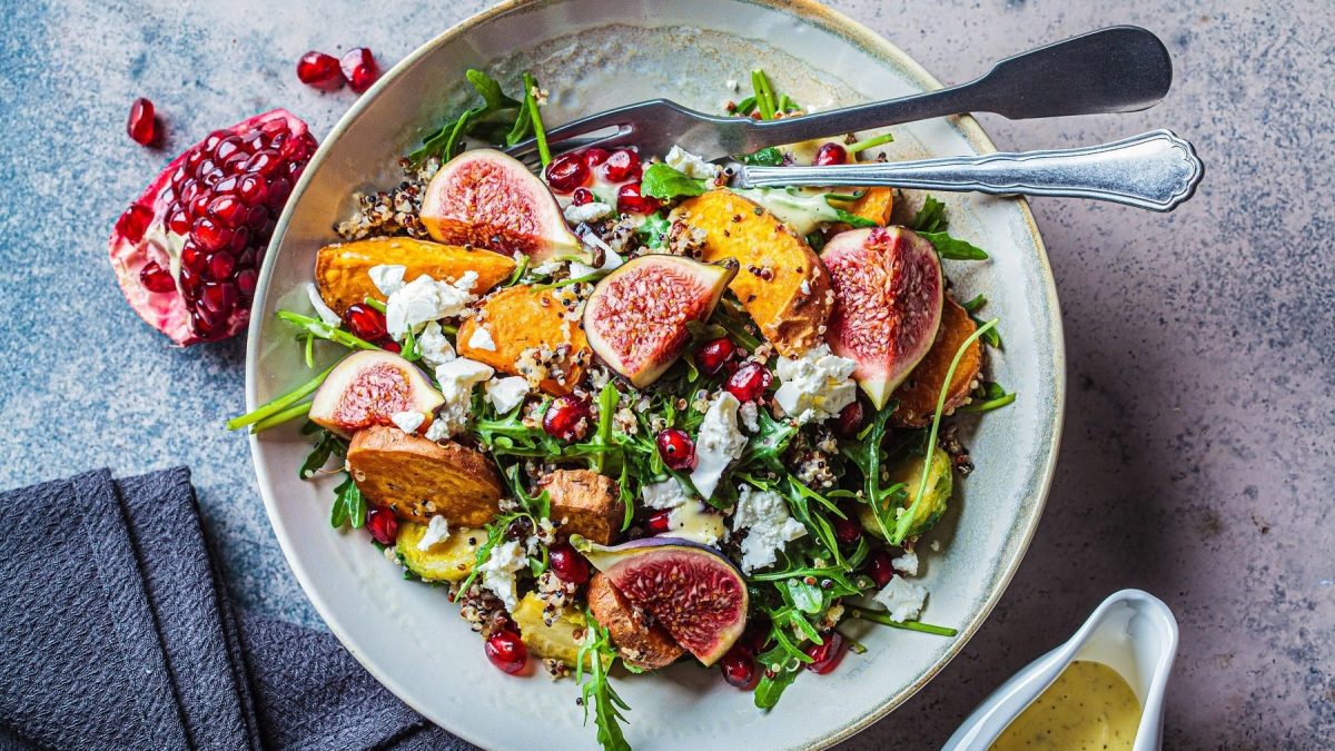 Quinoa-Salat mit Feigen, Süßkartoffelecken, Granatäpfeln, Feta und gebackenen Süßkartoffelecken, serviert in einer großen Keramikschale auf einem steinernen Tisch, angereicht mit einer cremigen Vinaigrette.