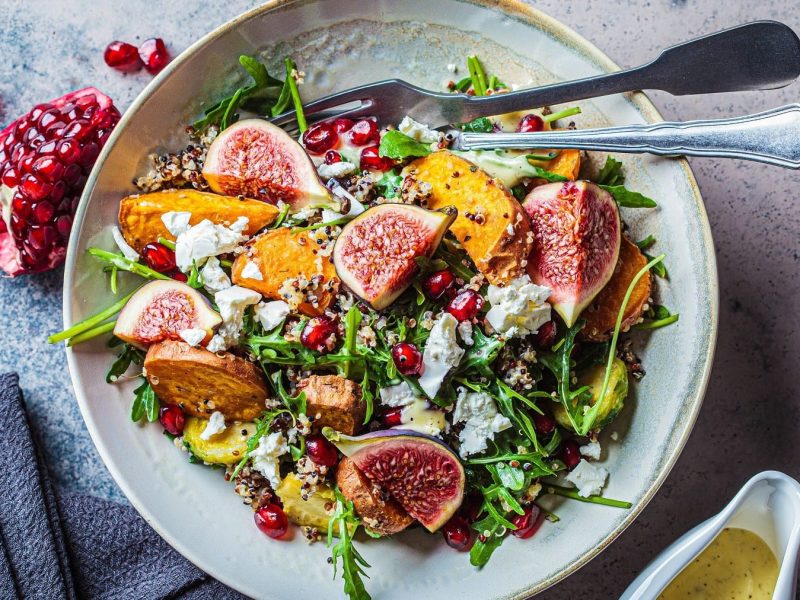 Quinoa-Salat mit Feigen, Süßkartoffelecken, Granatäpfeln, Feta und gebackenen Süßkartoffelecken, serviert in einer großen Keramikschale auf einem steinernen Tisch, angereicht mit einer cremigen Vinaigrette.