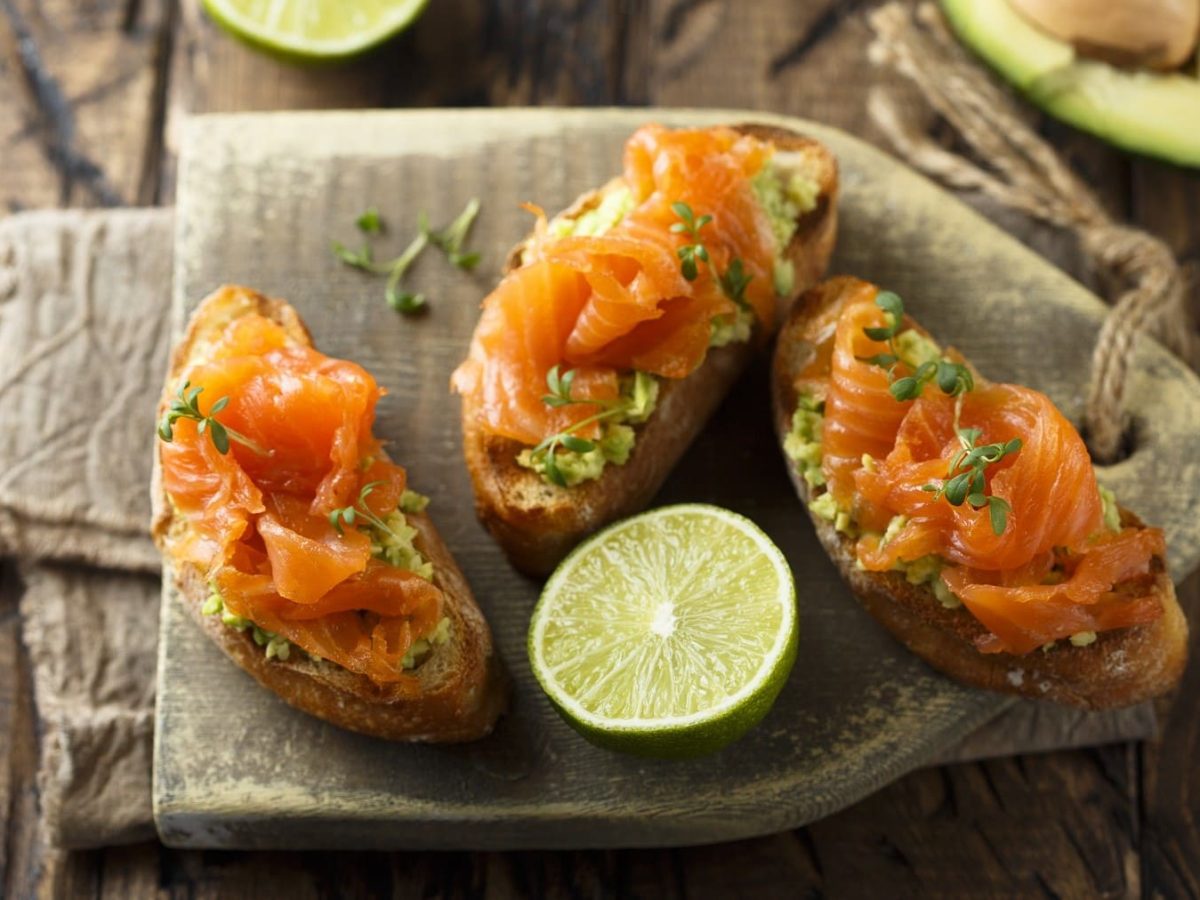 Avocado-Lachs-Bruschetta, garniert mit Limettensaft und frischer Kresse, serviert auf einem Holzbrett auf einem dunklen Holztisch.