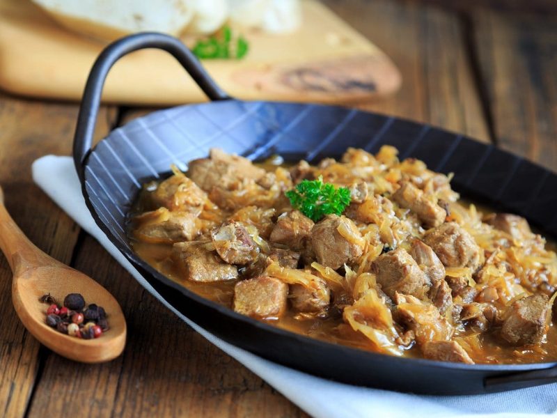 In einer rustikalen Pfanne wird original Szegediner Gulasch serviert. Neben der Pfanne liegen Pfefferkörner . Die Pfanne steht auf einem rustikalem Tisch.