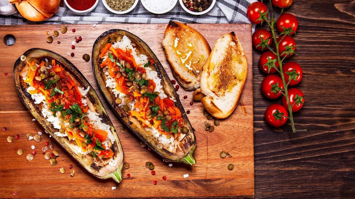 Gefüllte Aubergine mit Kürbis, Reis und Linsen serviert mit gebackenem Baguette auf einem Holzbrett.