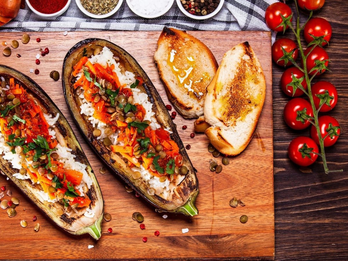 Gefüllte Aubergine mit Kürbis, Reis und Linsen serviert mit gebackenem Baguette auf einem Holzbrett.