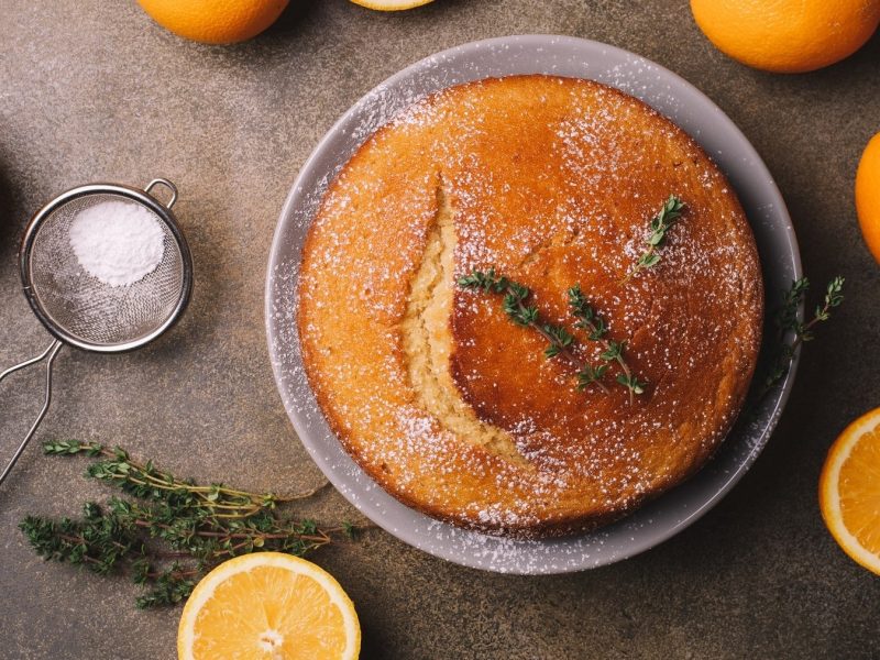 Getränkter Orangenkuchen, garniert mit Oregano und Puderzucker, serviert auf einer dunklen Steinplatte.