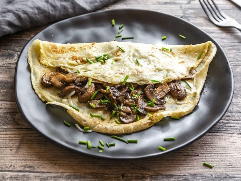 Deftige Pfannkuchen mit Champignons und Käsestreuseln gefüllt, serviert auf einem schwarzen Teller auf einem grauen Holztisch.