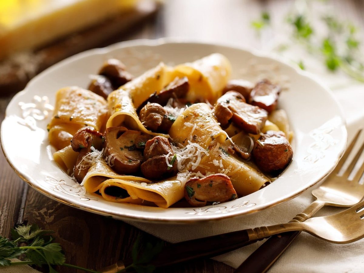 Auf einem rustikalem Holztisch steht ein weißer Teller mit Pappardelle mit Kraeutern und Pilzen.