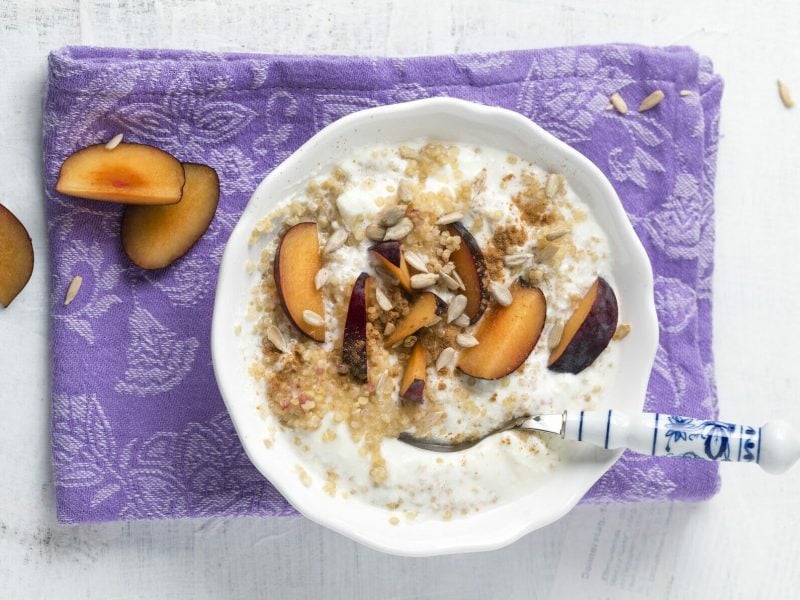 Pflaumen-Frühstücksbrei aus Quinoa mit Joghurt in weißer Schüssel auf lila Tuch