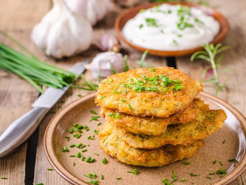 Kartoffelpuffer mit selbstgemachtem Quarkdip auf braunem Teller mit Schnittlauch, dahinter Knoblauch und Messer