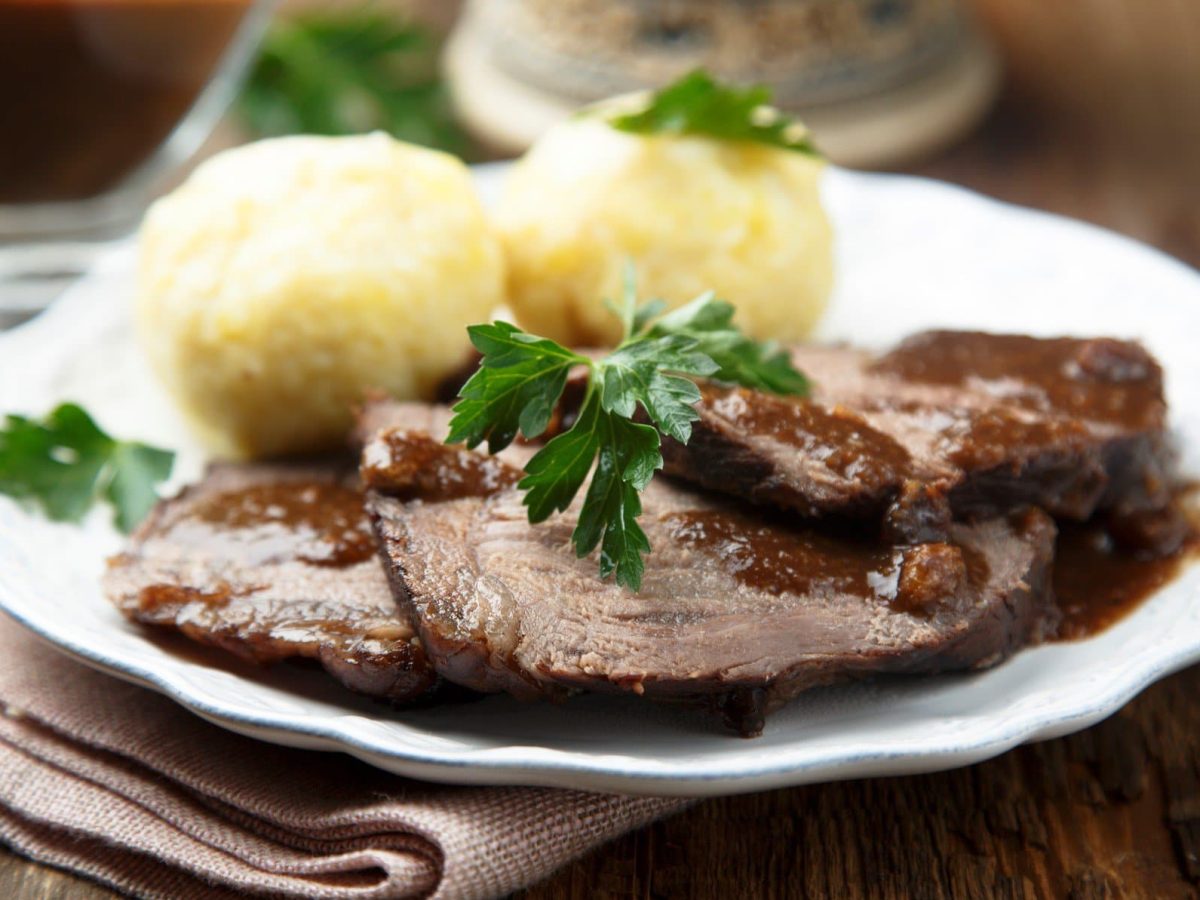 Sauerbraten aus Kalbsbäckchen mit Soße und Klößen auf weißem Teller, beigem Tuch und dunklem Tisch. Garniert mit Petersilie. Im Hintergrund Sauciere aus Glas mit Soße. Aufnahme aus der Frontalsicht.