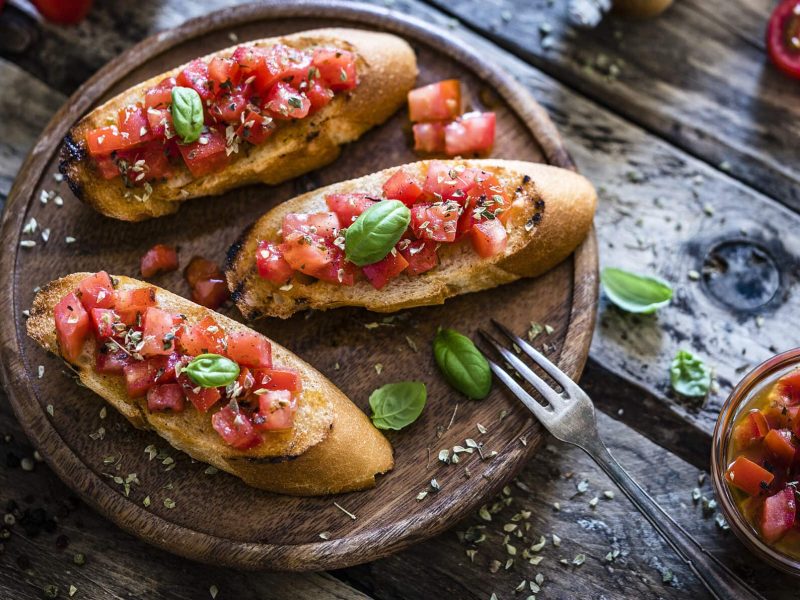 Einfache Tomaten-Bruschetta auf Holztablett, daneben Basilikum und Öl