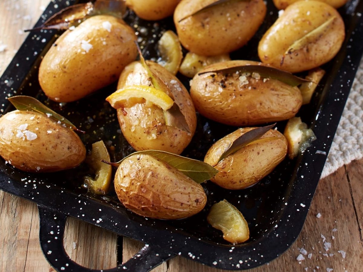 Auf einem Blech liegen Kartoffeln, in denen jeweils ein Lorbeer-Blatt steckt. Drumherum liegen Zitronenscheiben.