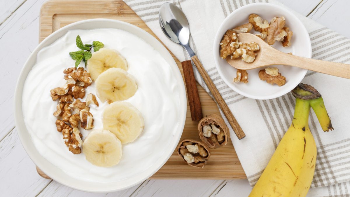 Quarkcreme mit karamellisierten Nüssen in weißer Schüssel vor Holzbrett, daneben Banane und Schüssel mit Walnüssen