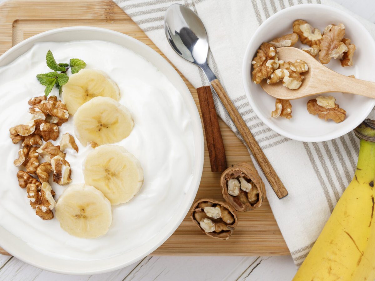 Quarkcreme mit karamellisierten Nüssen in weißer Schüssel vor Holzbrett, daneben Banane und Schüssel mit Walnüssen