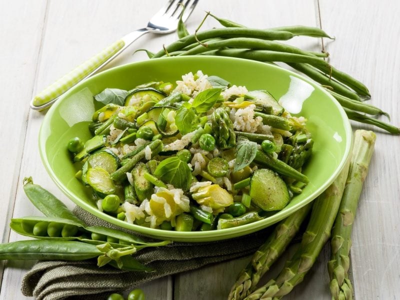 Grünes Spargel-Risotto mit Zucchini, Erbsen, Frühlingszwiebeln und frischem Basilikum, serviert in einer grünen Schale auf einem hellen Holztisch.