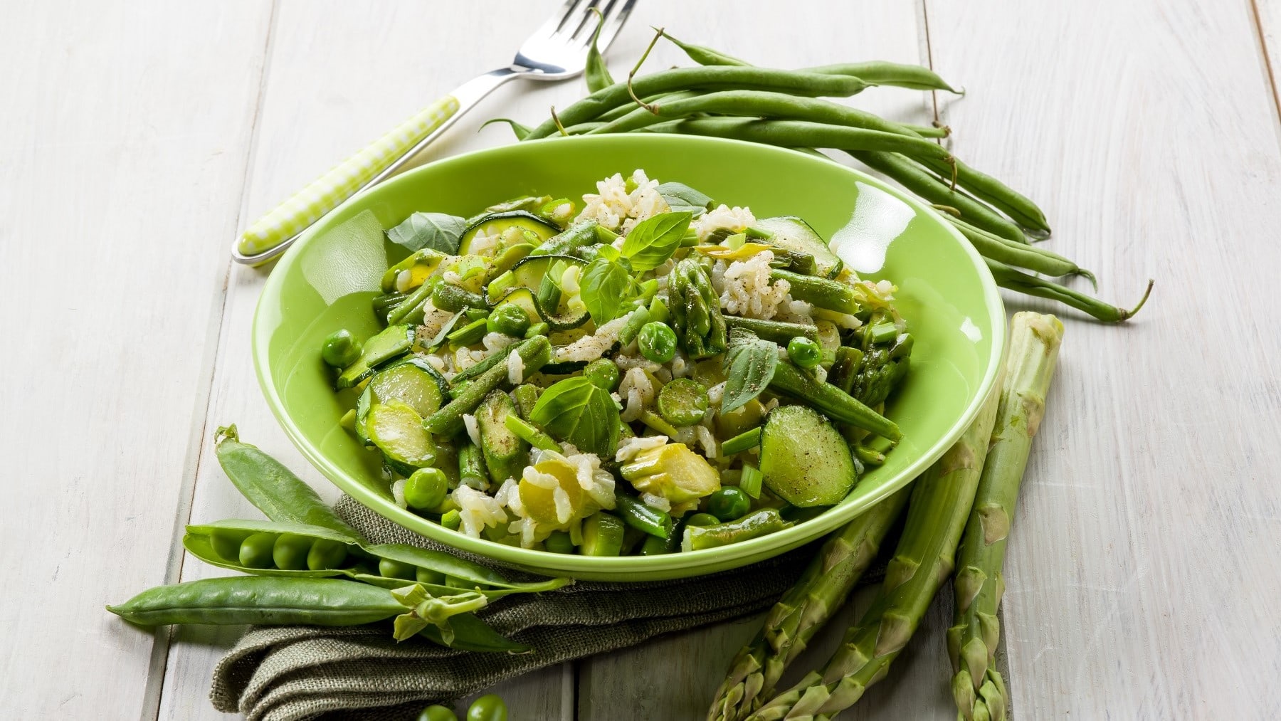 Grünes Spargel-Risotto mit Zucchini, Erbsen, Frühlingszwiebeln und frischem Basilikum, serviert in einer grünen Schale auf einem hellen Holztisch.
