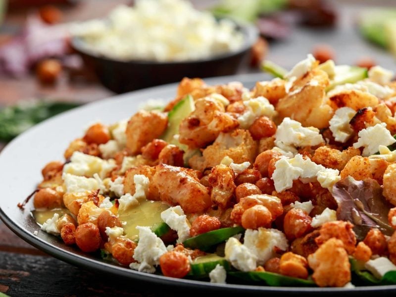 Salat mit geröstetem Blumenkohl, Gurke und Kichererbsen auf dunkler Platte, darüber Feta gebröselt