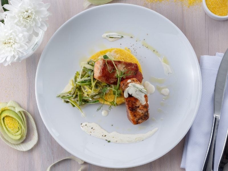 Gegrilltes Lachsfilet auf Polenta-Talern auf einem weißen Teller neben Besteck und Porree