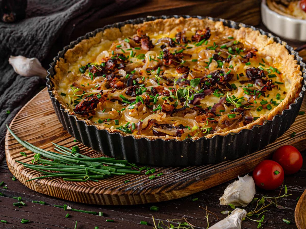 Vegetarischer Zwiebelkuchen mit Räuchertofu auf Holzuntergrund, daneben tomaten und Lräuter