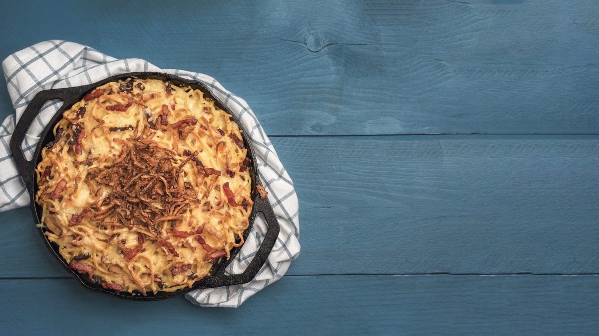 Kürbis-Käse-Spätzle, serviert mit Röstzwiebeln bestreut in einer großen, runden gusseisernen Form auf einem grauen Tisch.