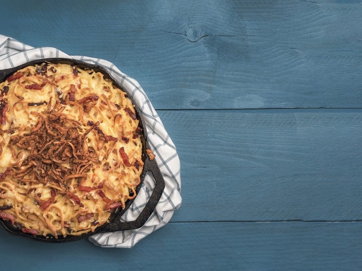 Kürbis-Käse-Spätzle, serviert mit Röstzwiebeln bestreut in einer großen, runden gusseisernen Form auf einem grauen Tisch.