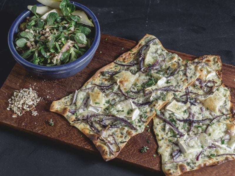 Flammkuchen mit Birne und Ziegenkäse auf Holzbrett, dahinter Salatschüssel