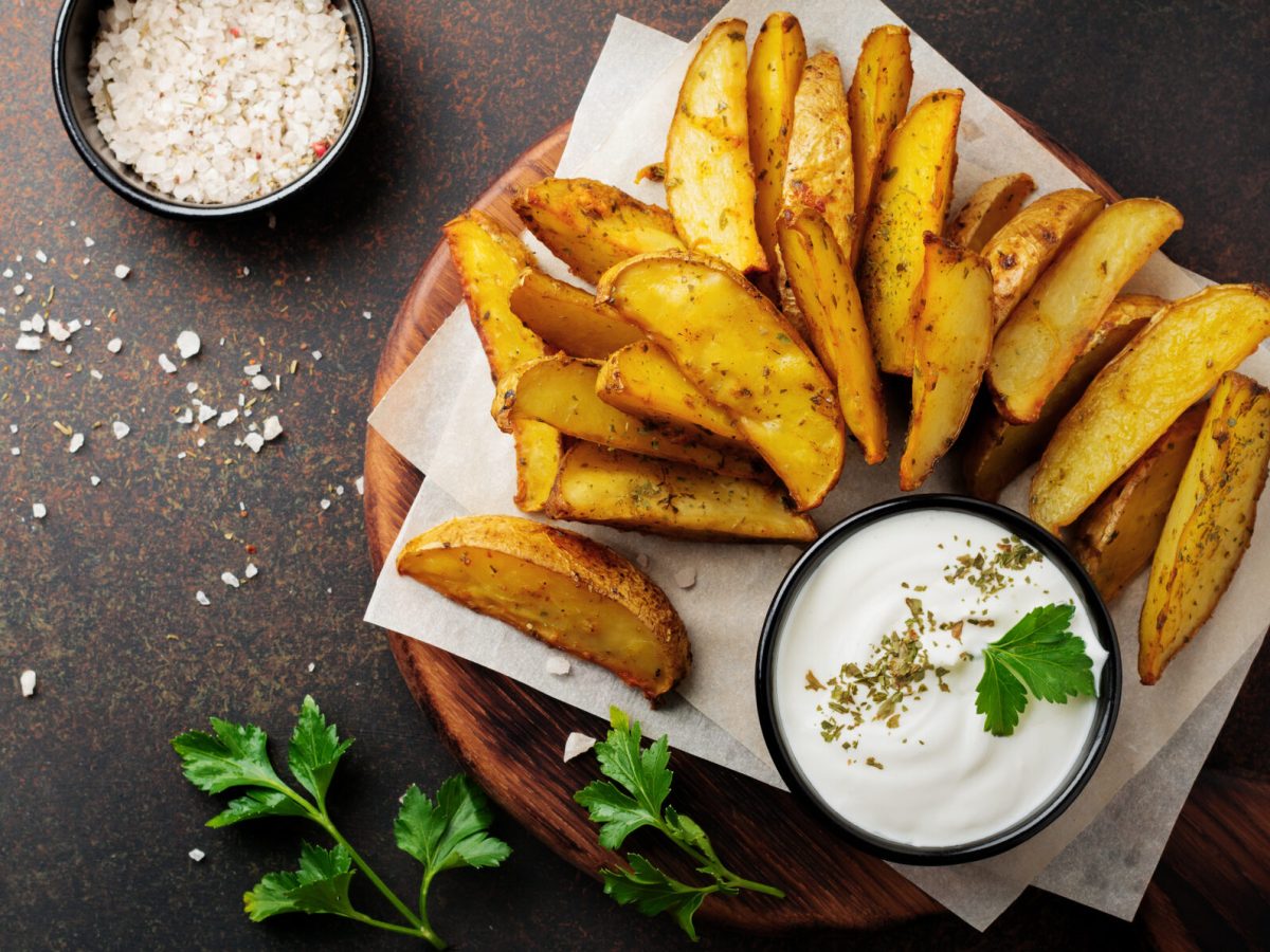 Kartoffel Wedges mit Skyr-Dip auf Papier, daneben Petersilie vor schwarzem Untergrund