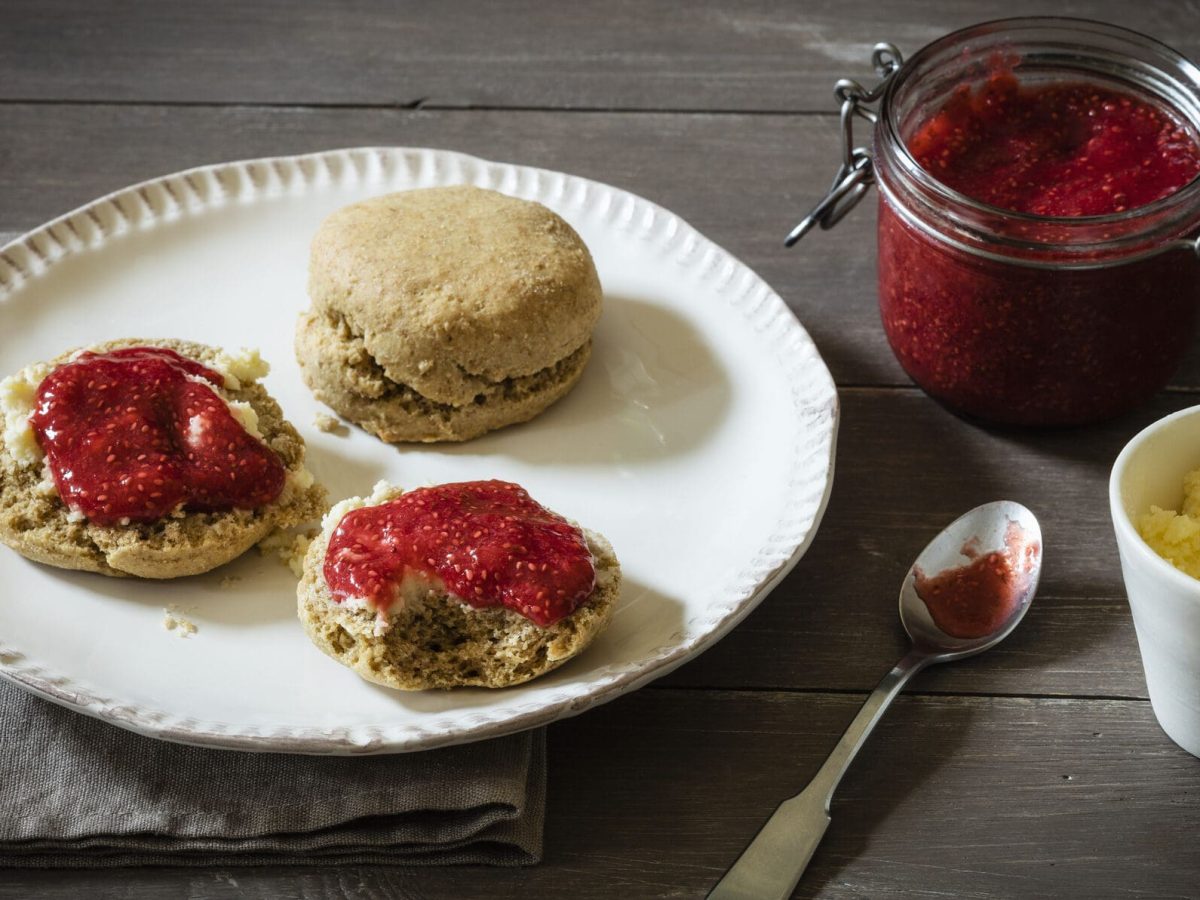 Britische Scones mit Erdbeermarmelade vor Holzuntergrund auf weißem Teller, daneben Marmelade und Clotted Cream