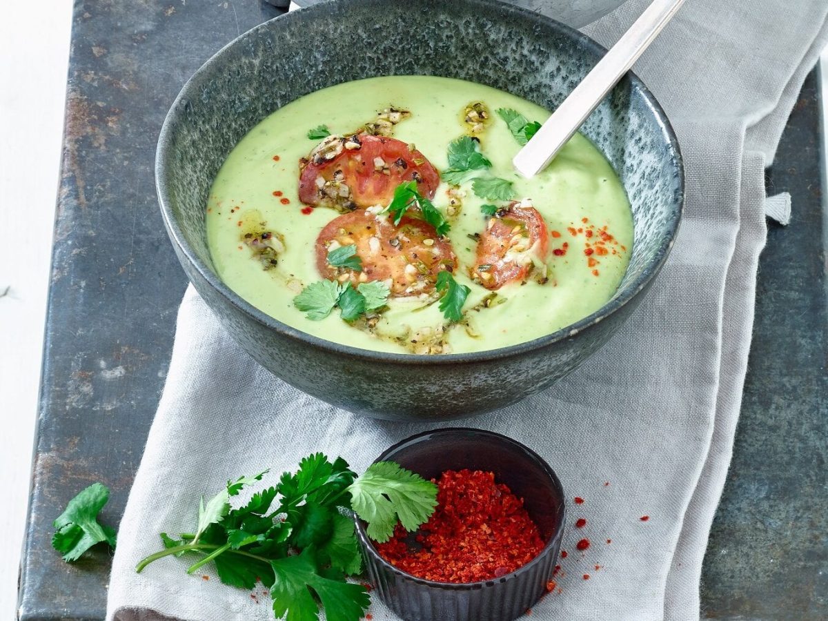 Kalte Avocado-Suppe mit getrockneten Tomaten in grauer Schüssel, daneben Chilliflocken