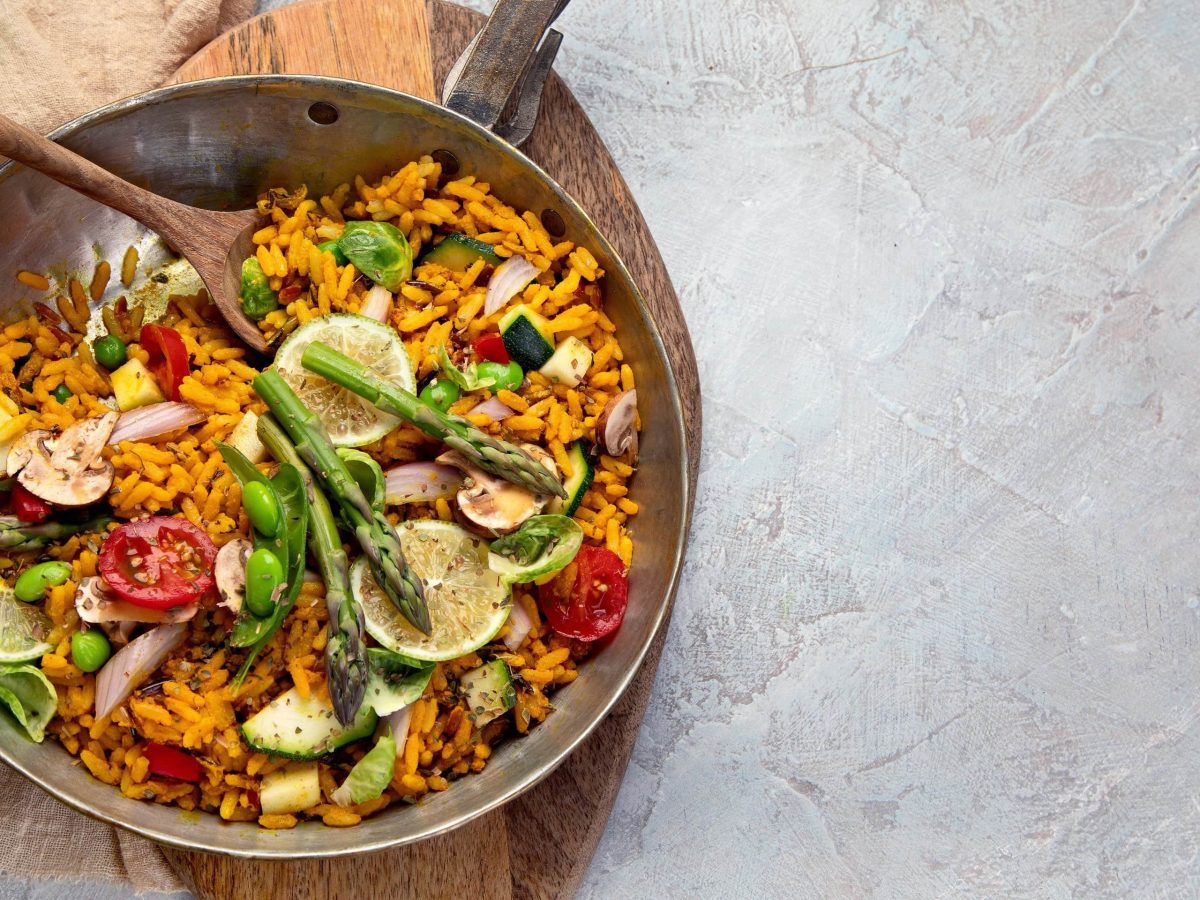 Bunte vegane Gemüse-Paella mit verschiedenen Gemüsesorten, garniert mit Zitronenhälften und Oregano in einer großen Kupferschale auf hellem Untergrund