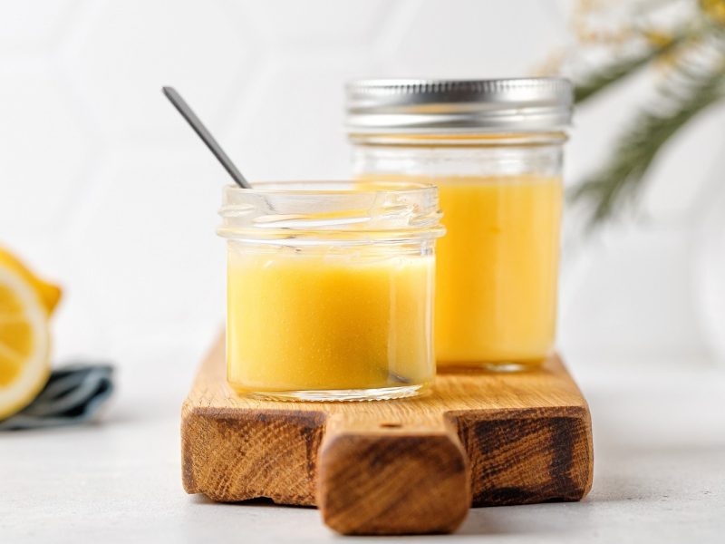 Selbst gemachtes Lemon Curd, abgefüllt in Einweck-Gläser und auf einem Holzbrett vor hellem Hintergrund serviert.