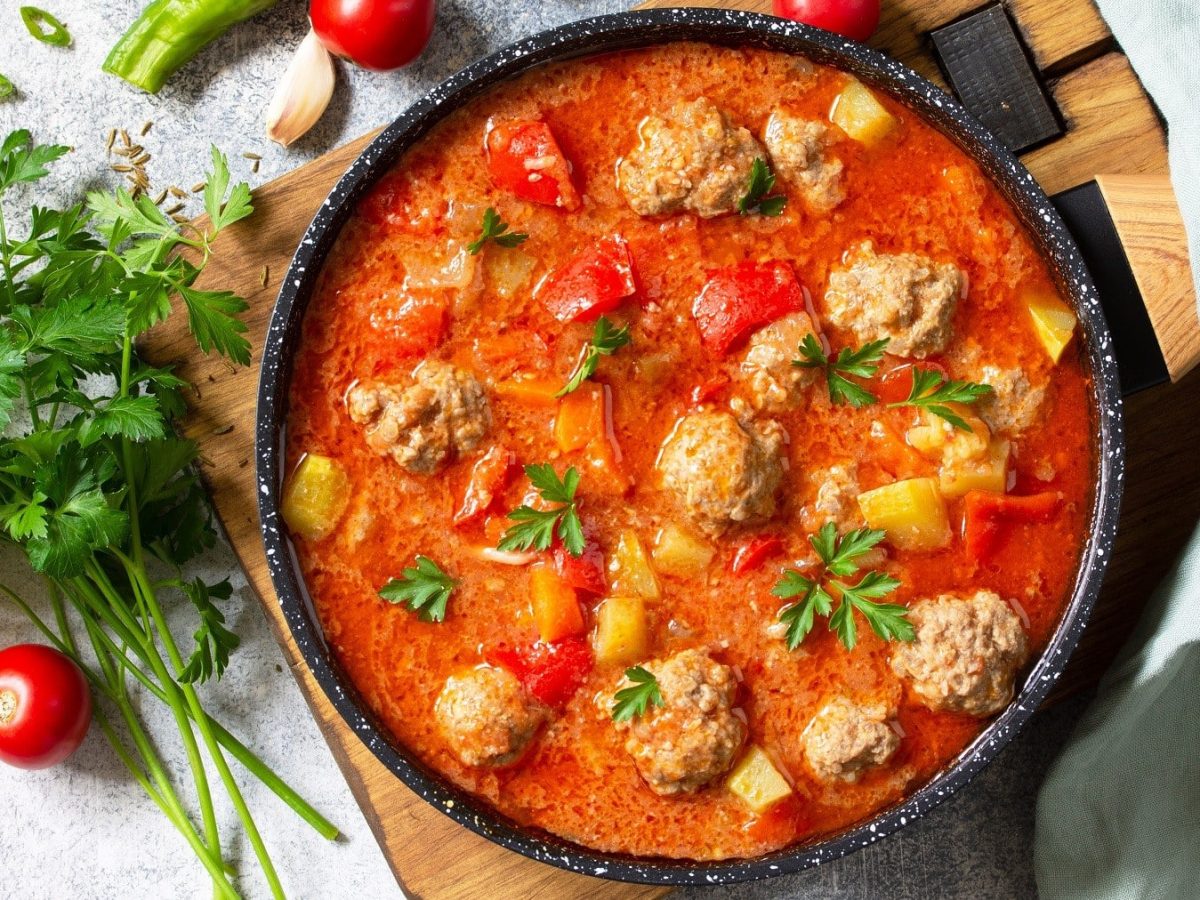 Linsen-Tomatensuppe mit Fleischbällchen, Paprika und frischer Petersilie, in einem großen Topf auf hellem Holzbrett serviert.