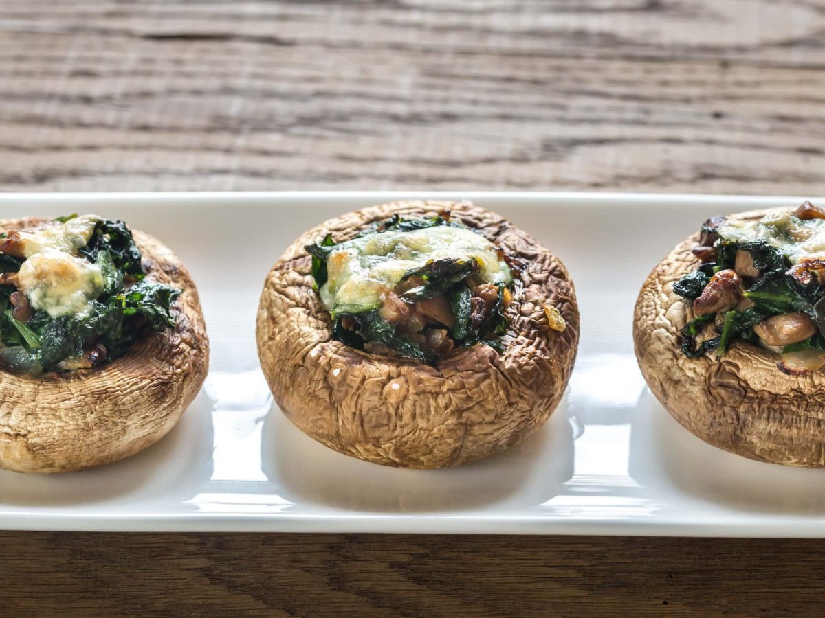 Mit Käse überbackene gefüllte Champignons mit Spinat, Speck und Zwiebeln, nebeneinander audgereiht auf einem weißen Teller auf einem hölzernen Tisch.