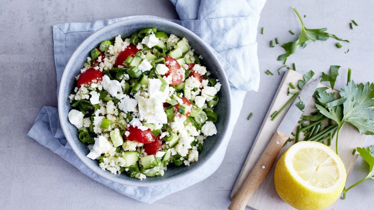 Couscous mit Feta, Petersilie, Gurke, Frühlingszwiebeln und Kirschtomaten in einer großen Schale, serviert auf einem Küchentuch auf einemhellen Tisch.