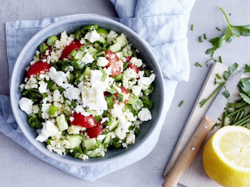 Couscous mit Feta, Petersilie, Gurke, Frühlingszwiebeln und Kirschtomaten in einer großen Schale, serviert auf einem Küchentuch auf einemhellen Tisch.