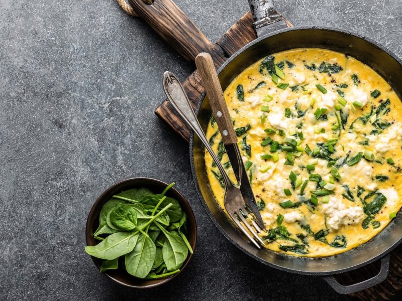 Aus der Vogelperspektive ist eine Pfanne zu sehen. IN der Pfanne ist ein Spinat-Feta-Omelett. Daneben liegen Messer und Gabel, sowie ein Schöächen mit Spinatblättern.