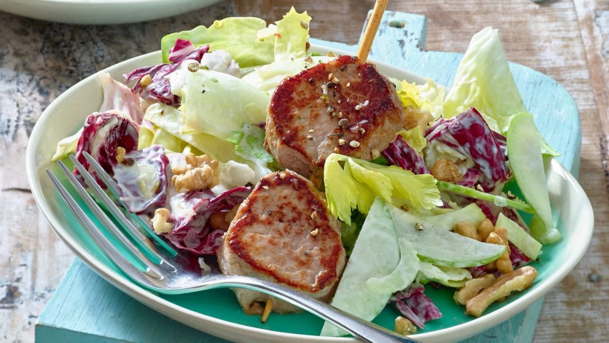 Holzuntergrund mit türkisblauer Servierte. Im Bildmittelpunkt ein Teller auf dem ein Waldorfsalat mit Schweinefilet angerichtet ist.