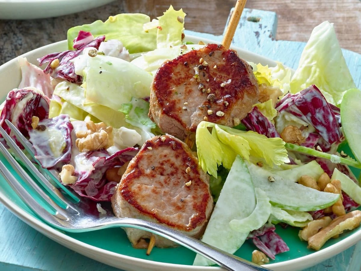 Holzuntergrund mit türkisblauer Servierte. Im Bildmittelpunkt ein Teller auf dem ein Waldorfsalat mit Schweinefilet angerichtet ist.