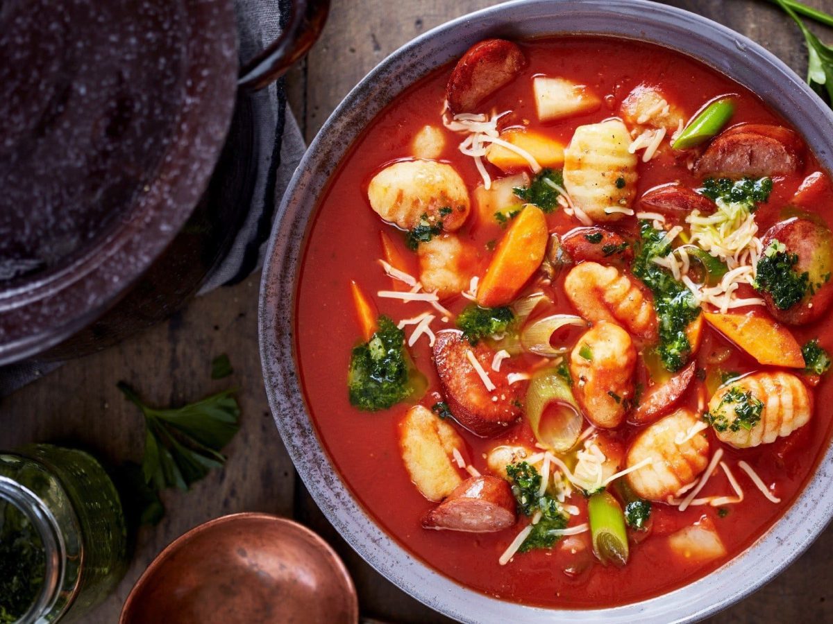 One-Pot-Gnocchi mit Wurst, Gemüse und Kräutern in einer deftigen Tomatensuppe als Eintopf, serviert in einer Tonschale auf hölzernem Untergrund