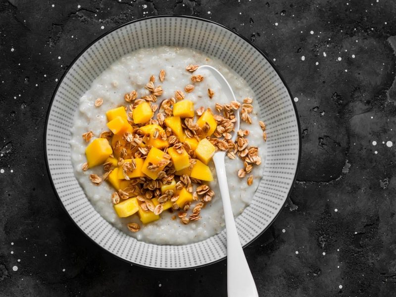 Haferbrei mit Kokos und Mangostückchen in einer Bowl, bestreut mit angerösteten Haferflocken und serviert auf einem dunklen Schiefertisch.