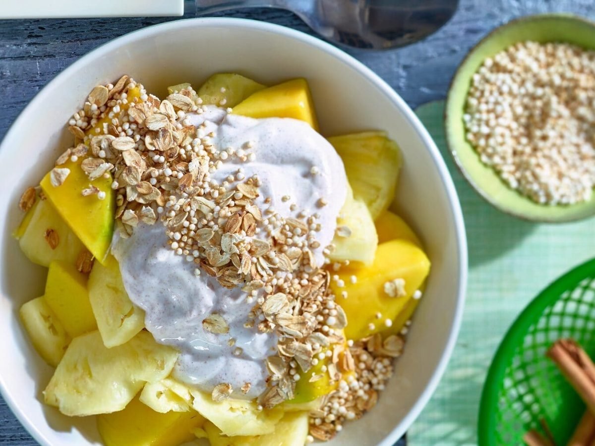 Eine Schale mit Ananas-Mango-Granola neben kleinen Schälchen mit Zutaten auf hellblauem Hintergrund.