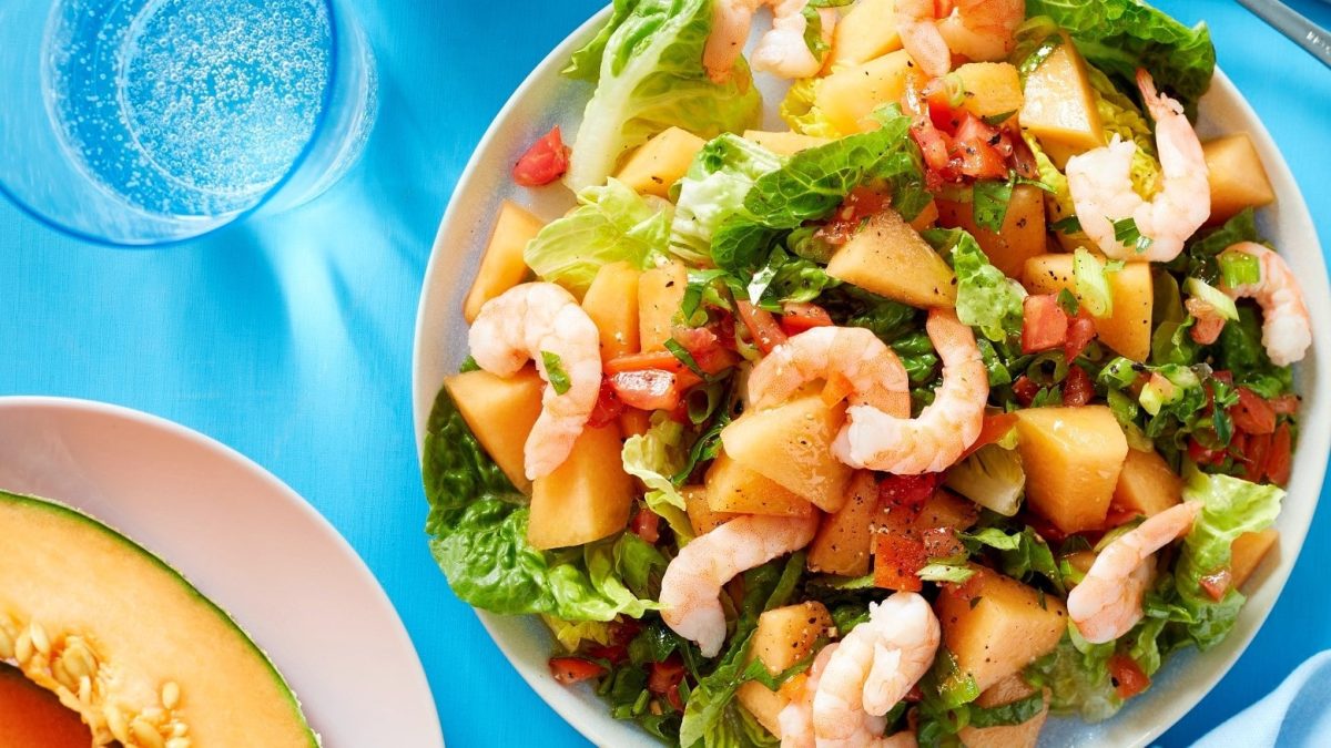 Ein heller Teller mit dem Melonen-Garnelen Salat. Oberhalb Messer und Gabel und ein Glas Wasser. Unten im Bild der Rand eines weiteren Tellers mit einer angeschnittenen Melone auf einer blauen Tischdecke.