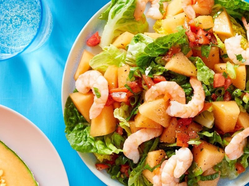 Ein heller Teller mit dem Melonen-Garnelen Salat. Oberhalb Messer und Gabel und ein Glas Wasser. Unten im Bild der Rand eines weiteren Tellers mit einer angeschnittenen Melone auf einer blauen Tischdecke.