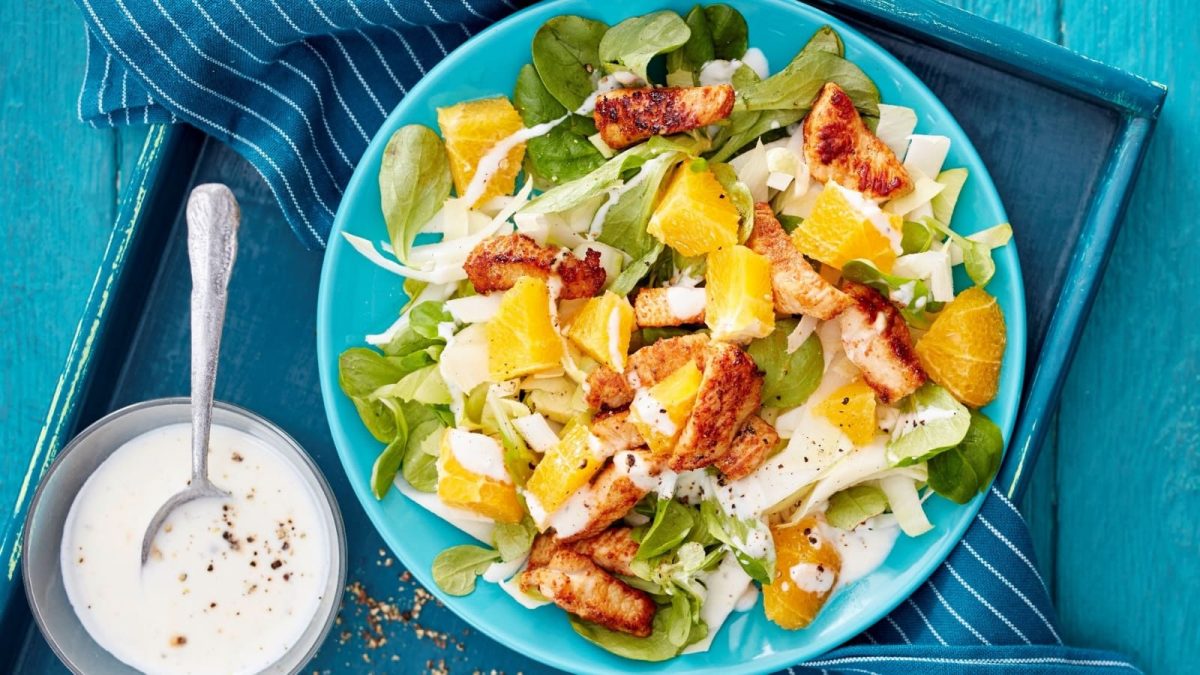 Draufsicht: Türkiser Holztisch mit blauem Tablett auf dem ein türkiser Teller mit Chicorée-Puten-Salat mit Orangen steht. Links daneben ein kleines Schälchen mit Dressing.