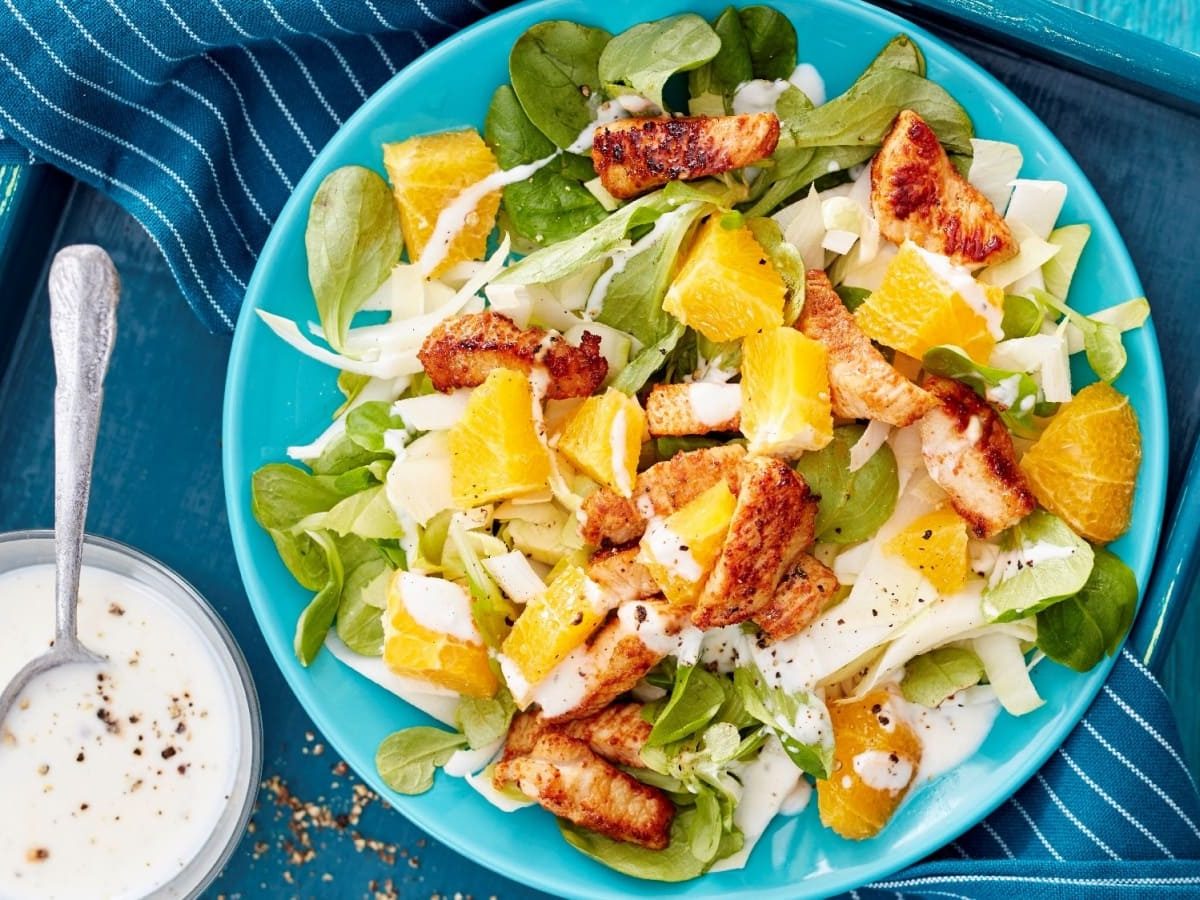 Draufsicht: Türkiser Holztisch mit blauem Tablett auf dem ein türkiser Teller mit Chicorée-Puten-Salat mit Orangen steht. Links daneben ein kleines Schälchen mit Dressing.