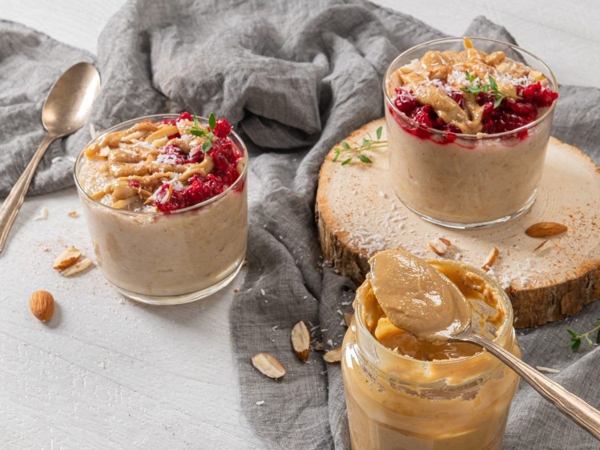 Porridge mit Himbeeren und Mandelmus, Kokosflocken und frischem Oregano garniert, serviert in Dessertschälchen auf einem grauen Tuch.