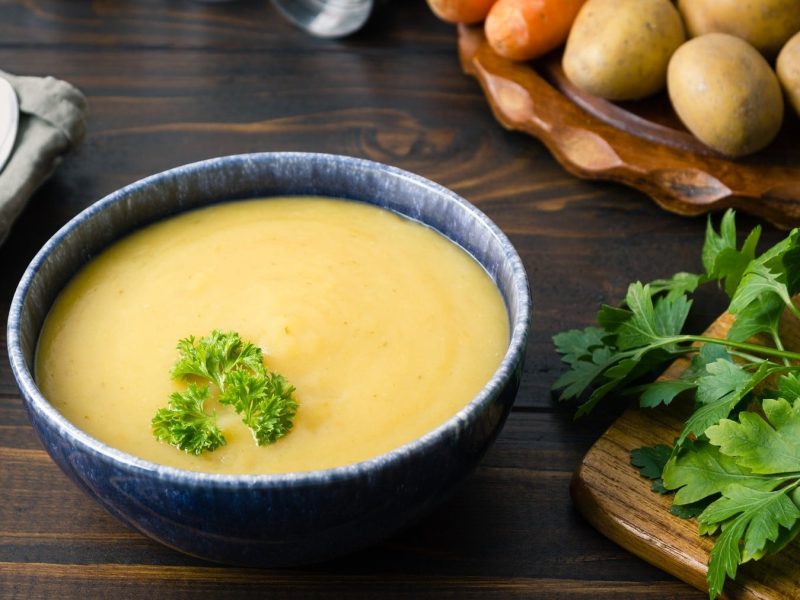 Pürierte Kartoffel-Cremesuppe mit Wurzelgemüse und garniert mit frischer Petersilie in einer dunklen Schale auf einem rustikalen Tisch.