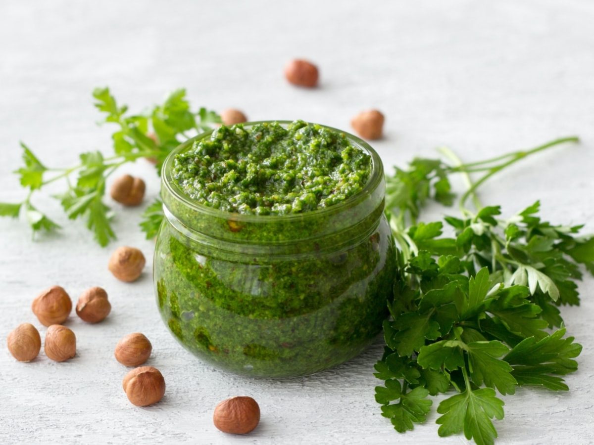 Hellgrauer Untergrund mit einem Glas im Bildmittelpunkt. Gefüllt mit selbst gemachtem Haselnuss-Petersilien-Pesto. Dekoriert mit frischer Petersilie und Haselnüssen.