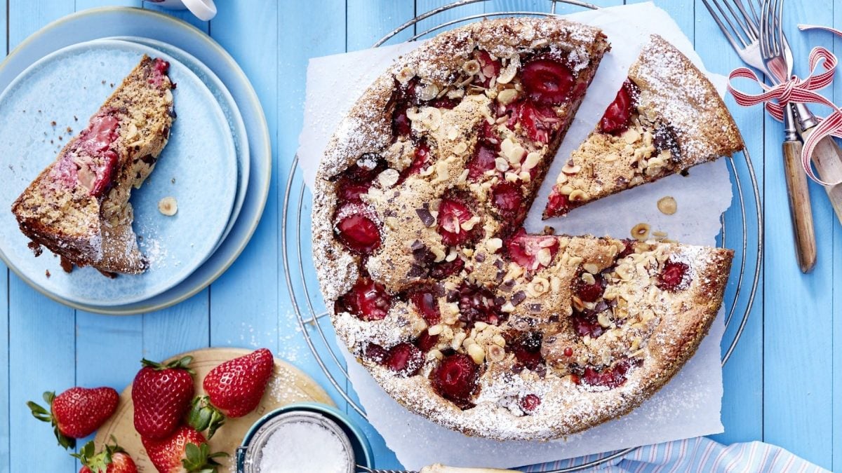 Saftiger, versunkener Erdbeerkuchen mit Nüssen und Schokoladensplittern, angeschnitten serviert auf einem hellblauen Holztisch, garniert mit Puderzucker.