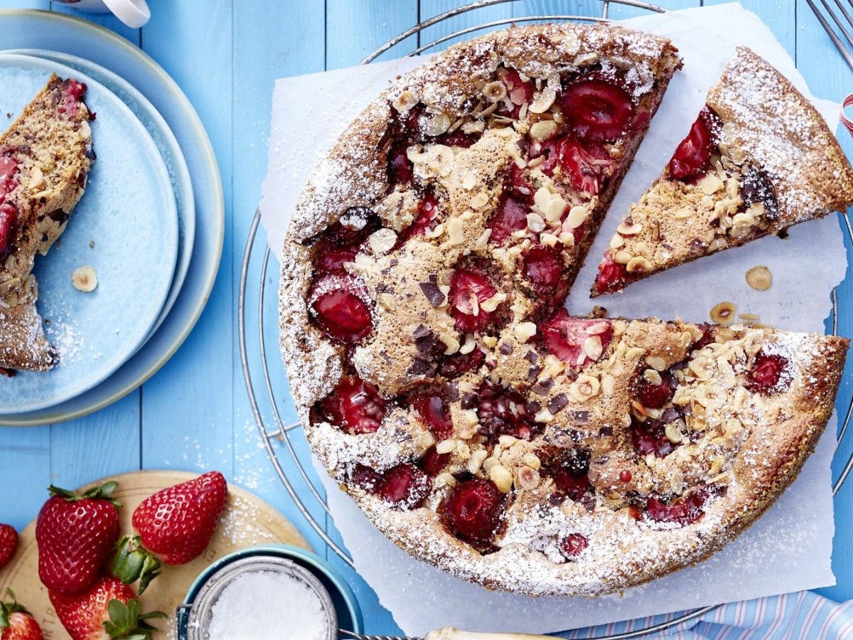 Saftiger, versunkener Erdbeerkuchen mit Nüssen und Schokoladensplittern, angeschnitten serviert auf einem hellblauen Holztisch, garniert mit Puderzucker.