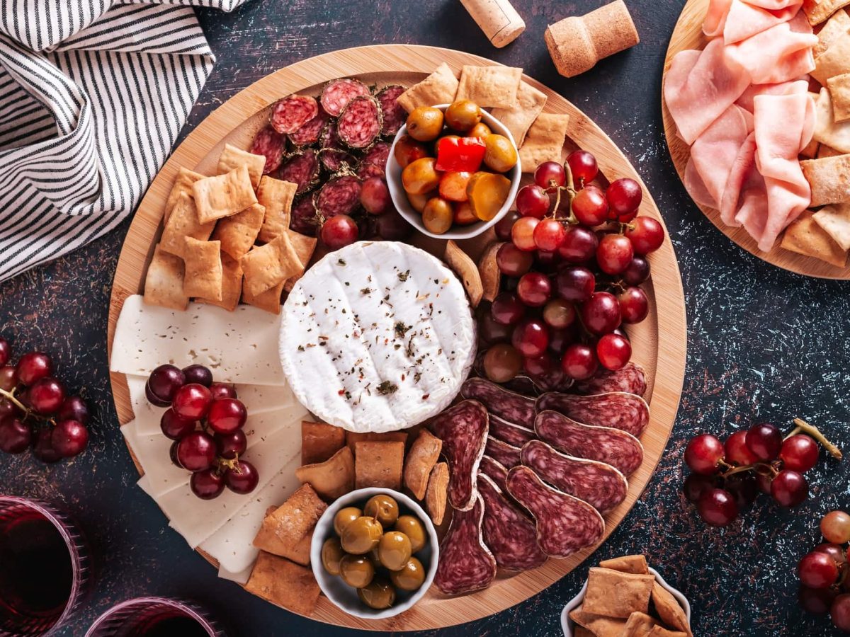 Ein rundes Holzbrett auf einem Marmortisch. Das Brett ist üppig belegt mit verschiedenen Wurst- und Käsesorten, Weintrauben, Oliven und Crackern. Daneben ein weiteres Brett mit Schinken und Crackern, außerdem sind auf dem Tisch Weintrauben, ein Geschirrtuch und Weingläser.